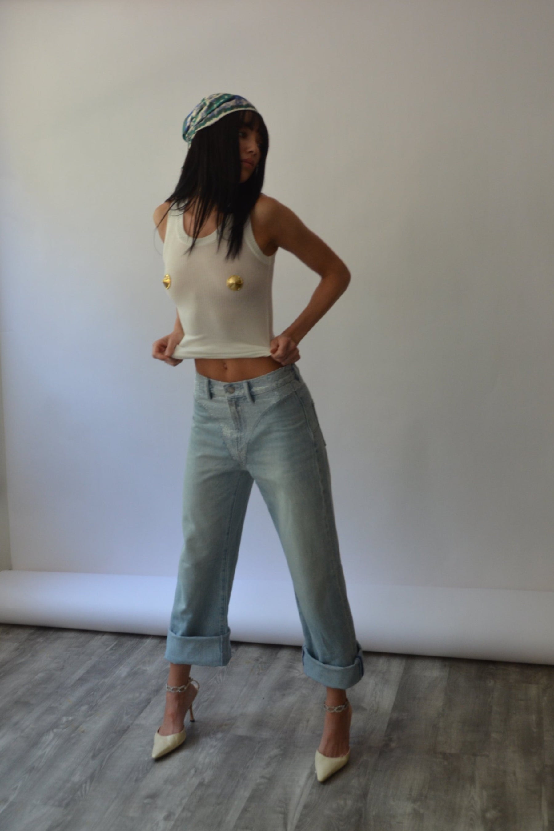 Person wearing a white shirt with gold pasties and light blue jeans standing against a white wall.