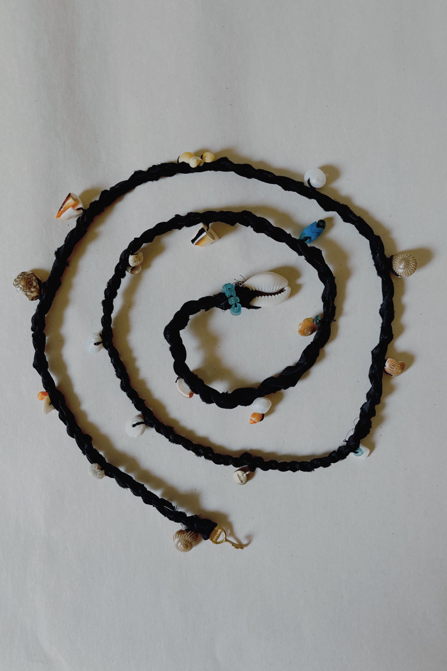 A black silk rope adorned with natural shells, beads, and pearls, displayed on a white background.