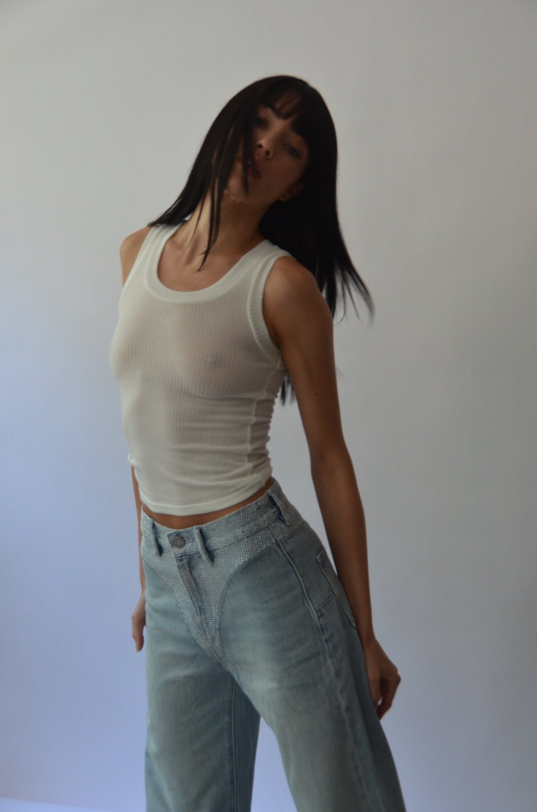 Person wearing a white tank top and blue jeans against a plain background