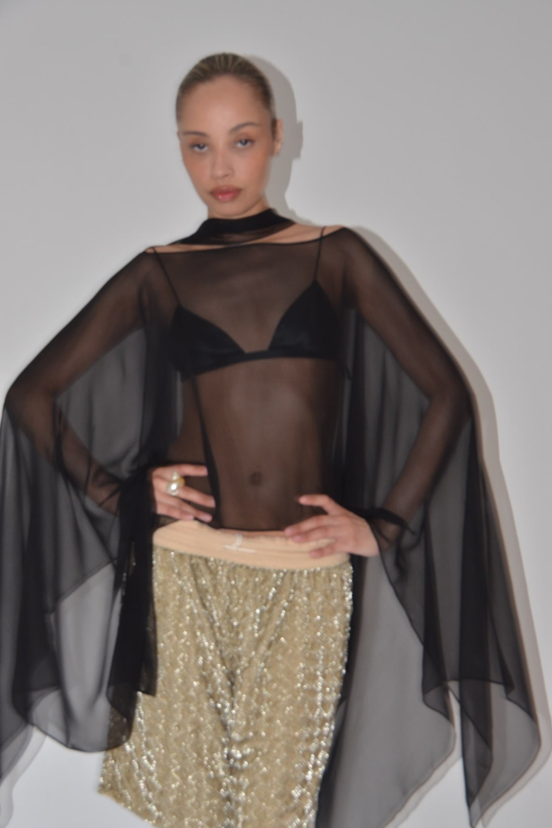Model wearing a sheer black top with a gold skirt on a reflective surface.