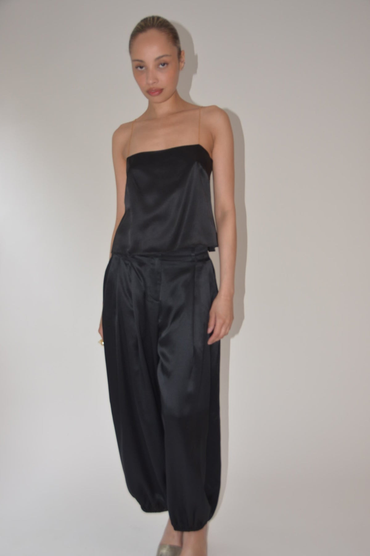 A woman modeling a black satin strapless top and matching wide-leg harem pants against a plain light gray studio background.