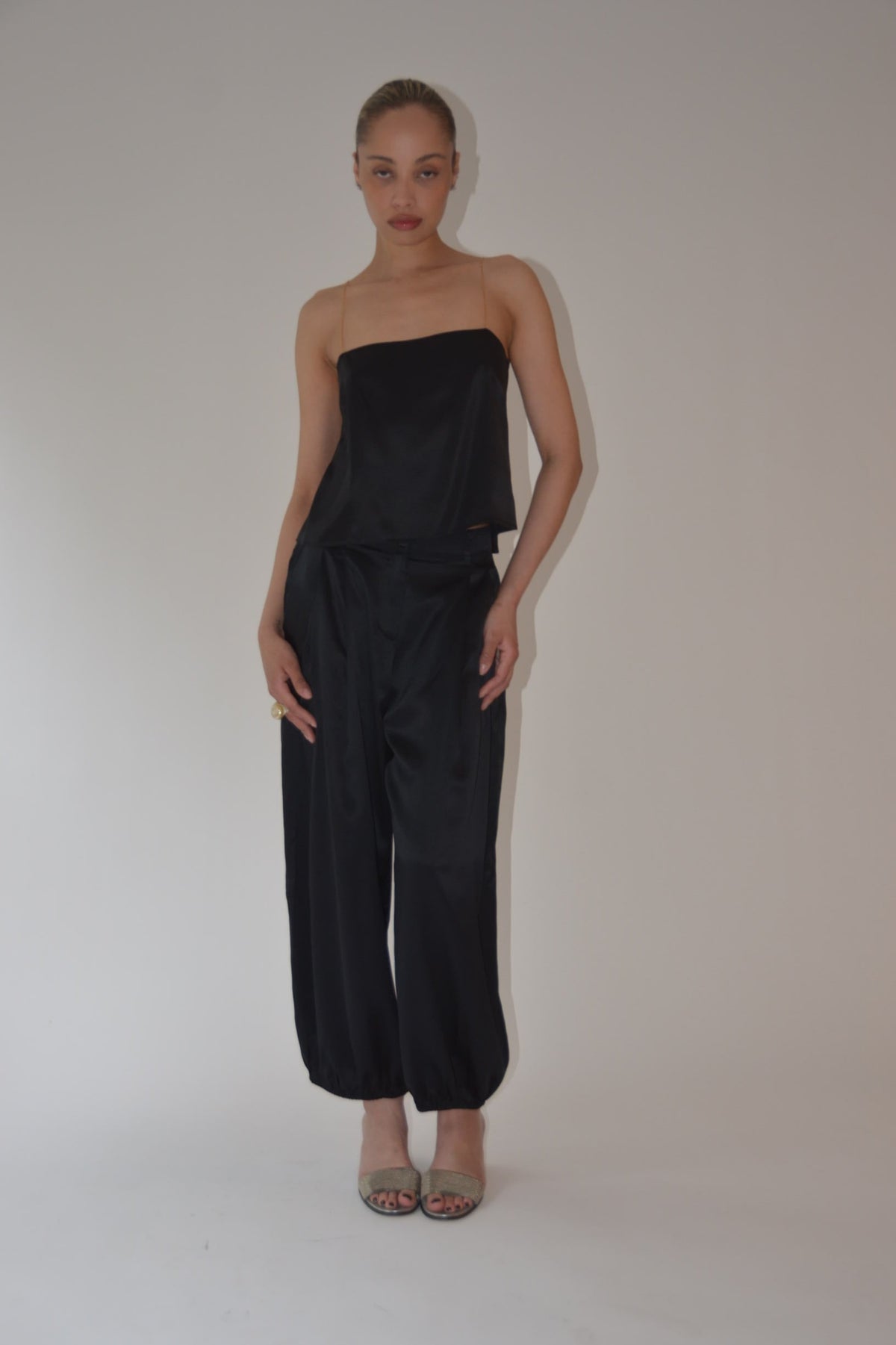 A woman modeling a black satin strapless top and matching wide-leg pants set, standing against a plain light gray studio background.
