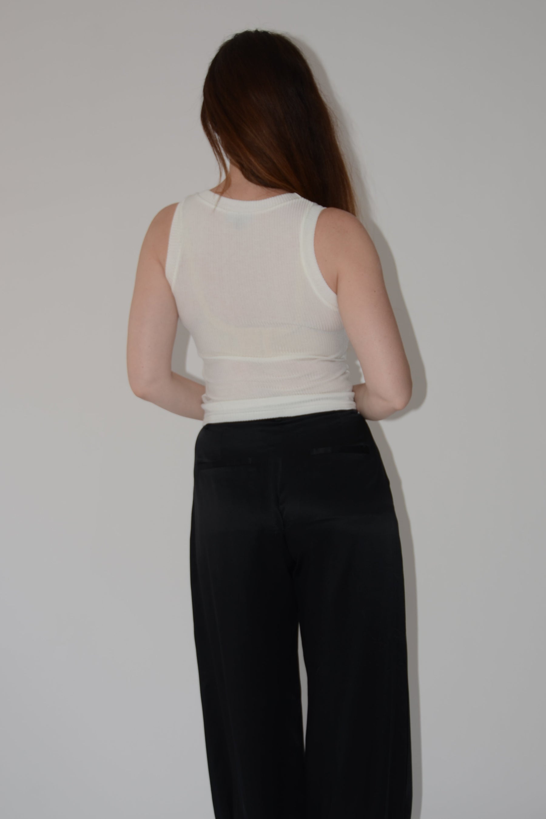 A woman viewed from behind wearing a white sleeveless knit top and black wide-leg pants, standing against a plain light gray wall.