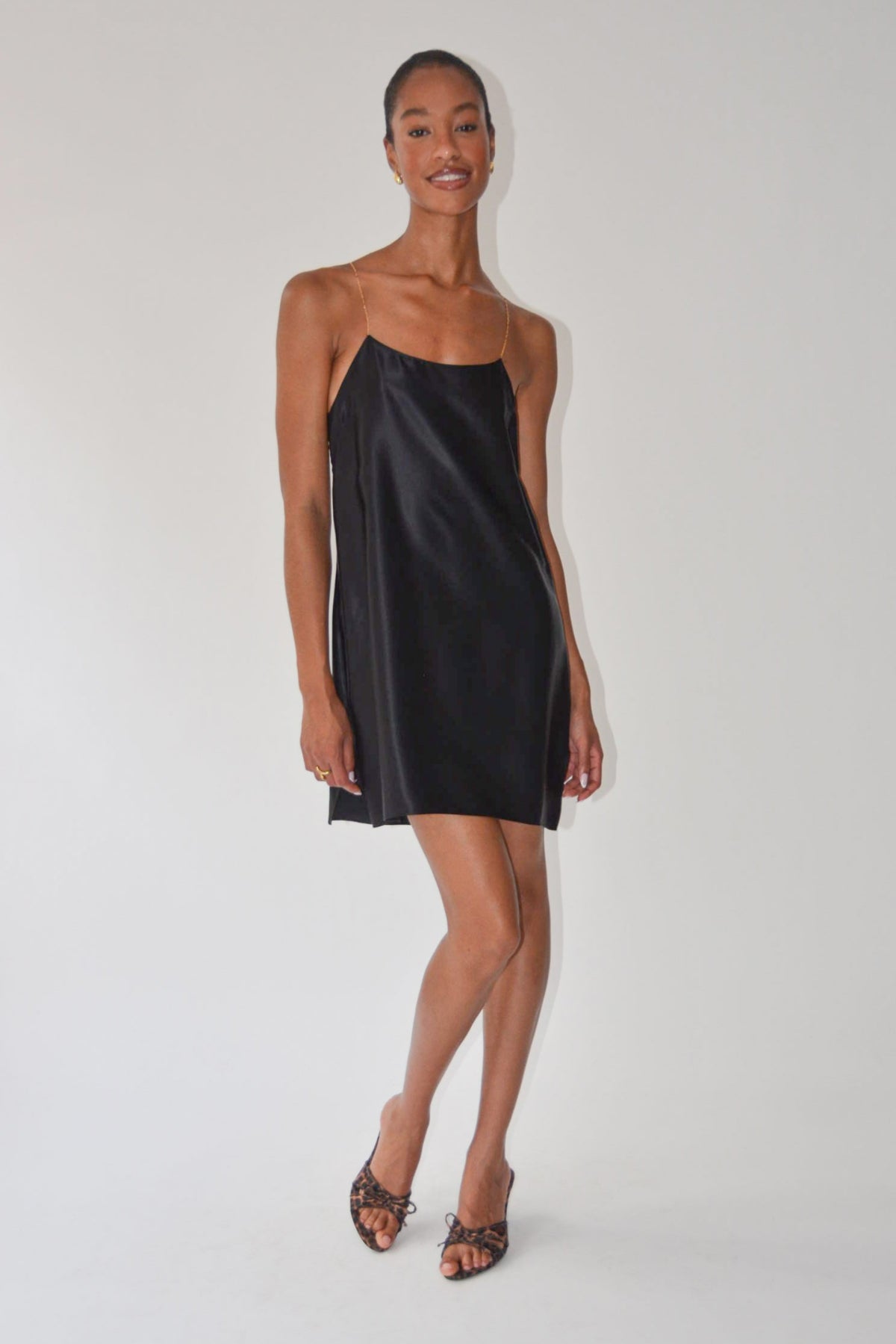 A woman modeling a black satin slip dress with a scoop neckline and spaghetti straps, paired with brown strappy heels, against a plain light gray studio background.