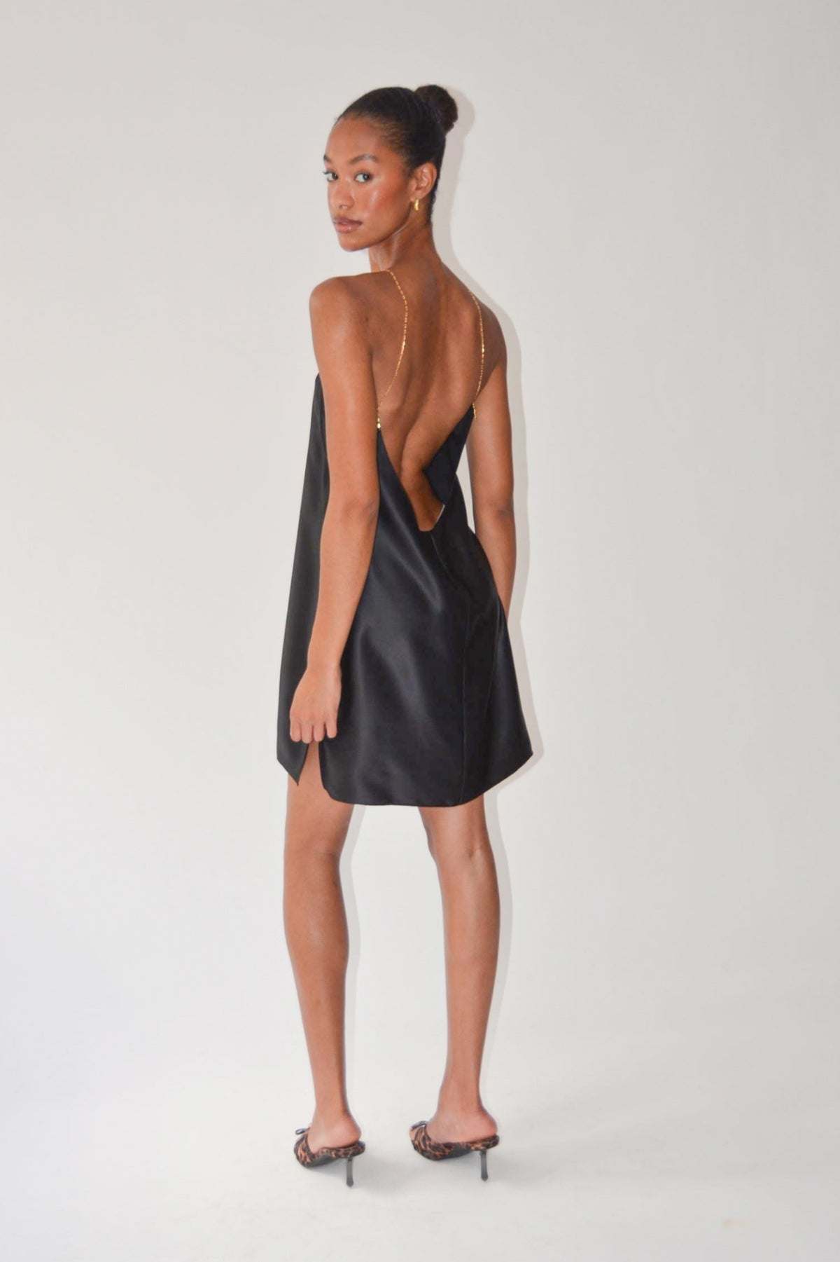 A woman modeling a black satin slip dress with a low open back, shown from the back against a plain white studio background.