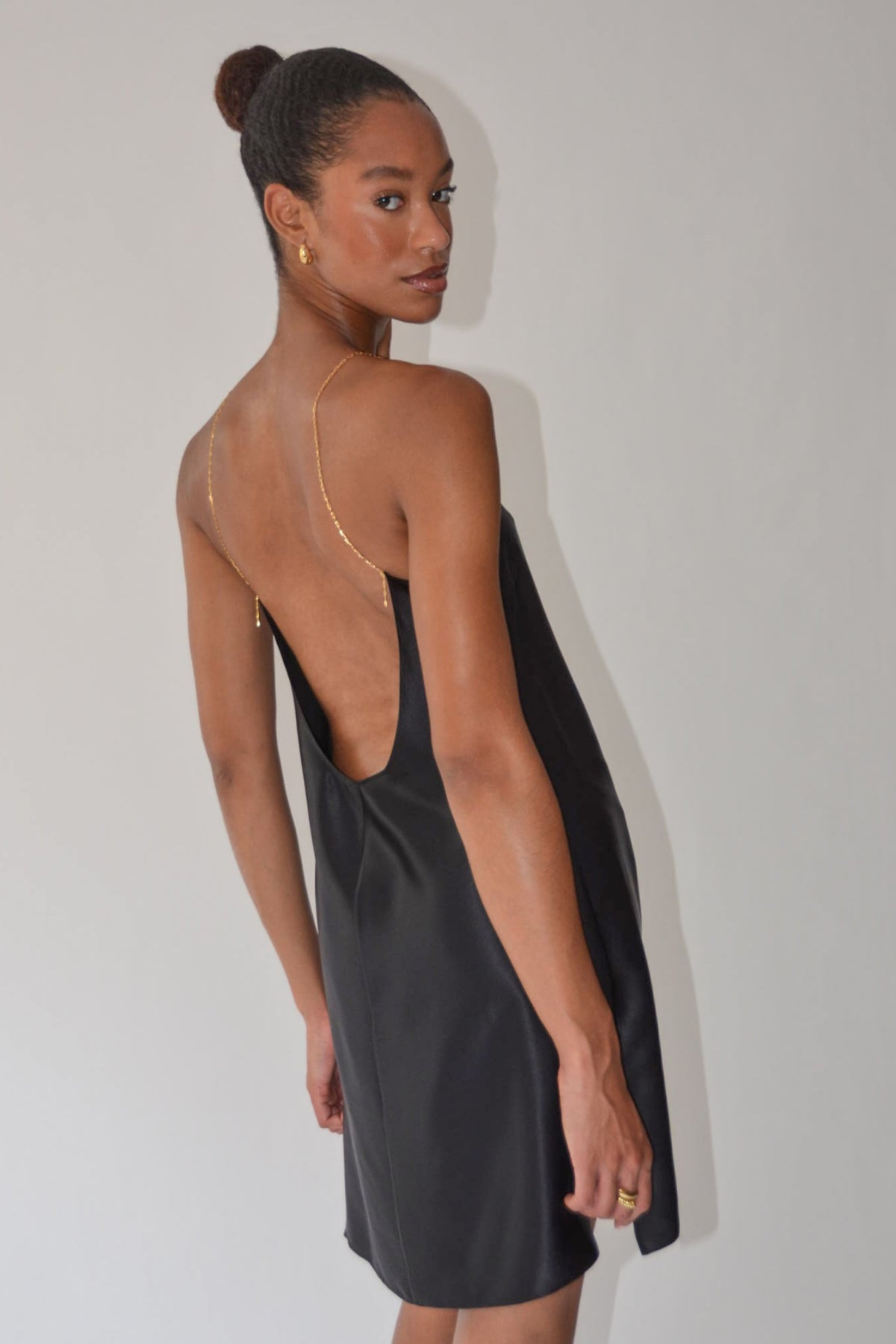 A woman wearing a black satin slip dress with a deep open back, photographed from behind against a plain light gray studio background.
