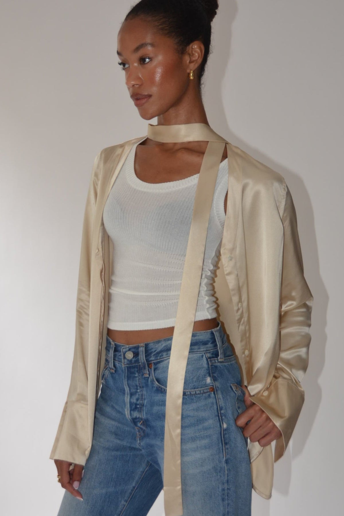 A model wearing a champagne gold satin jacket with a matching ribbon choker, styled over a white ribbed crop top and blue jeans, standing against a plain light gray wall.