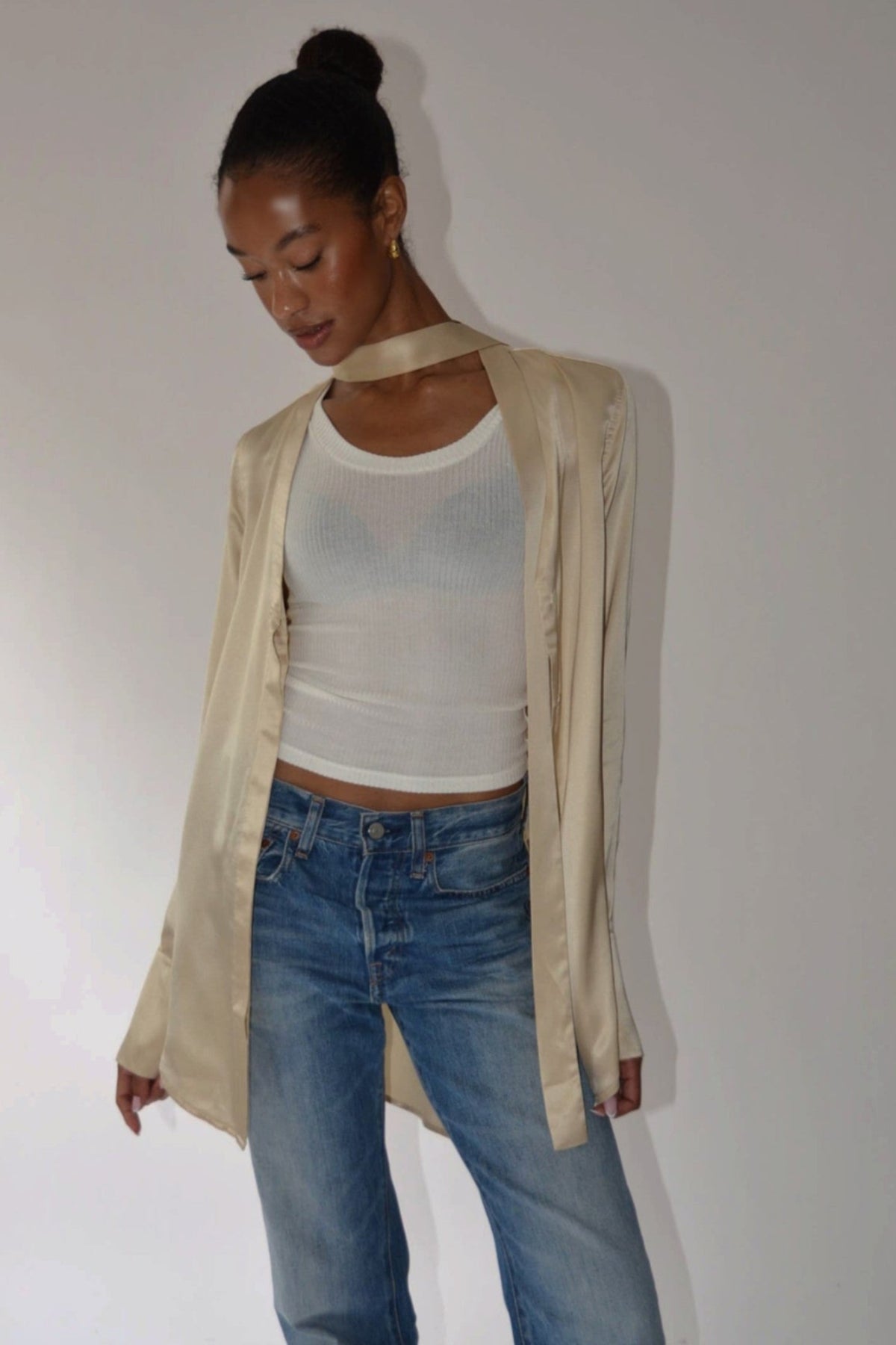 A woman wearing a beige satin-like open-front jacket over a white ribbed tank top and blue jeans, standing against a plain light gray wall.