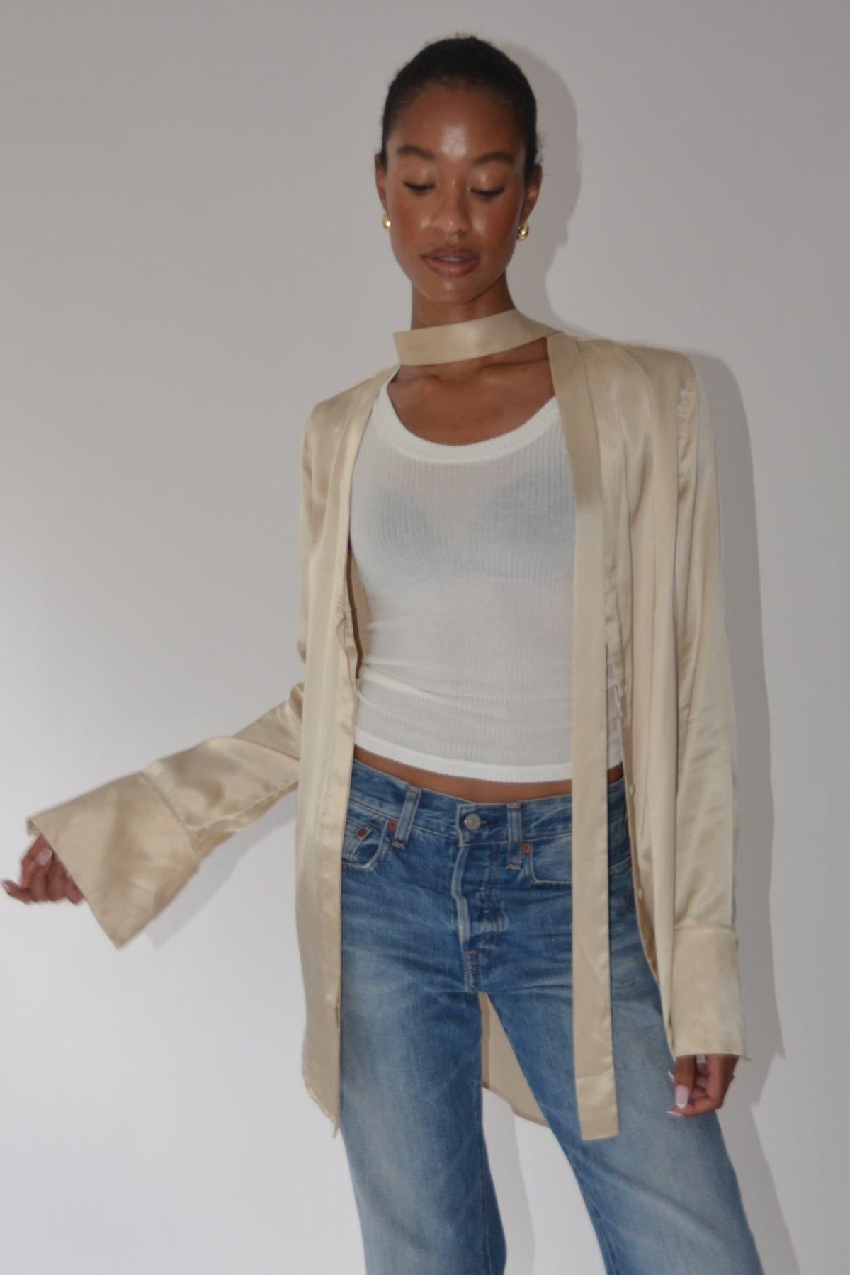 A model wearing a beige open-front long-sleeve jacket with a high collar, layered over a white tank top and blue jeans, standing against a plain light gray wall.