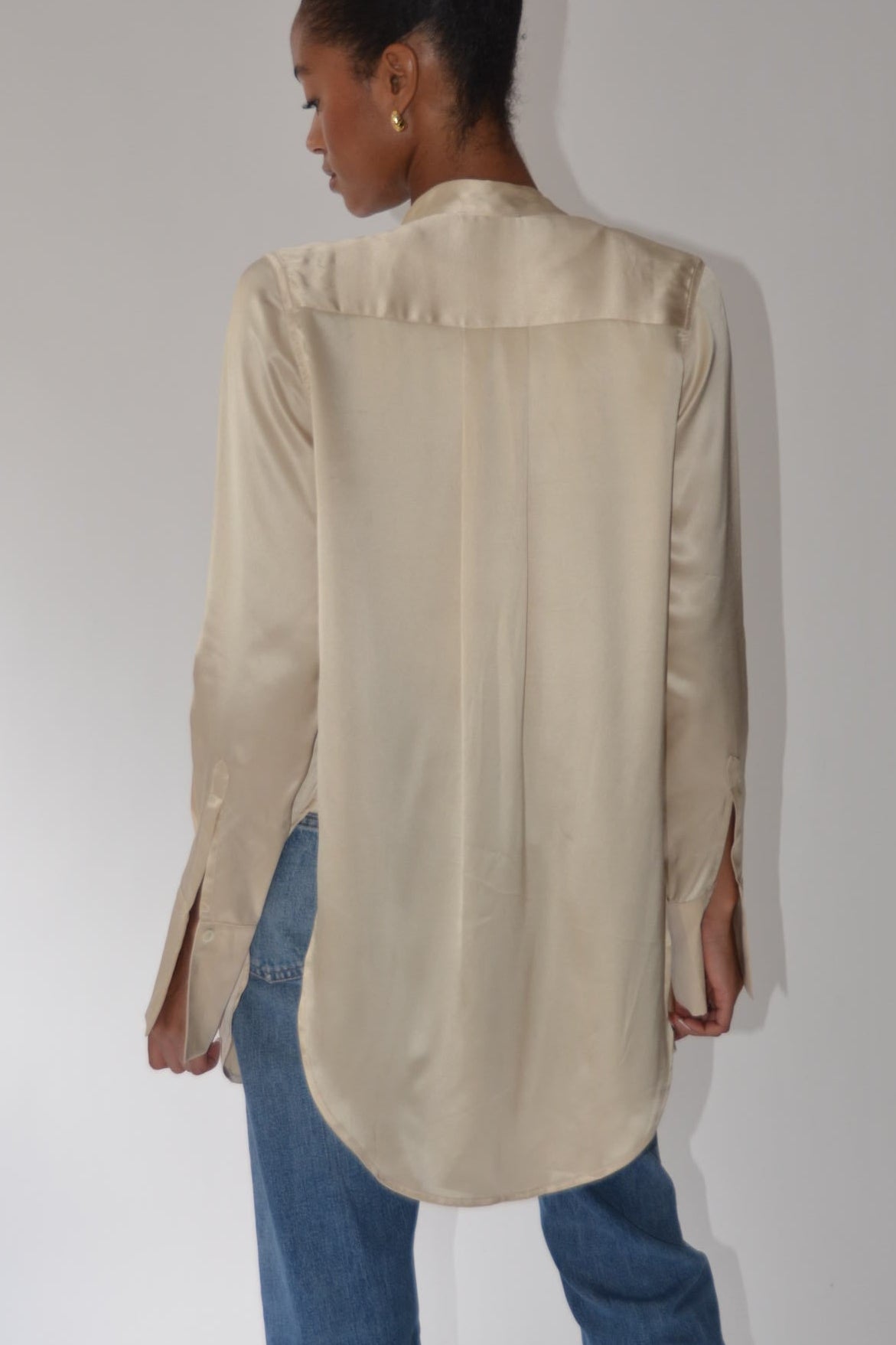A woman viewed from the back wearing a light beige satin long-sleeve blouse and blue jeans, standing against a plain white studio background.