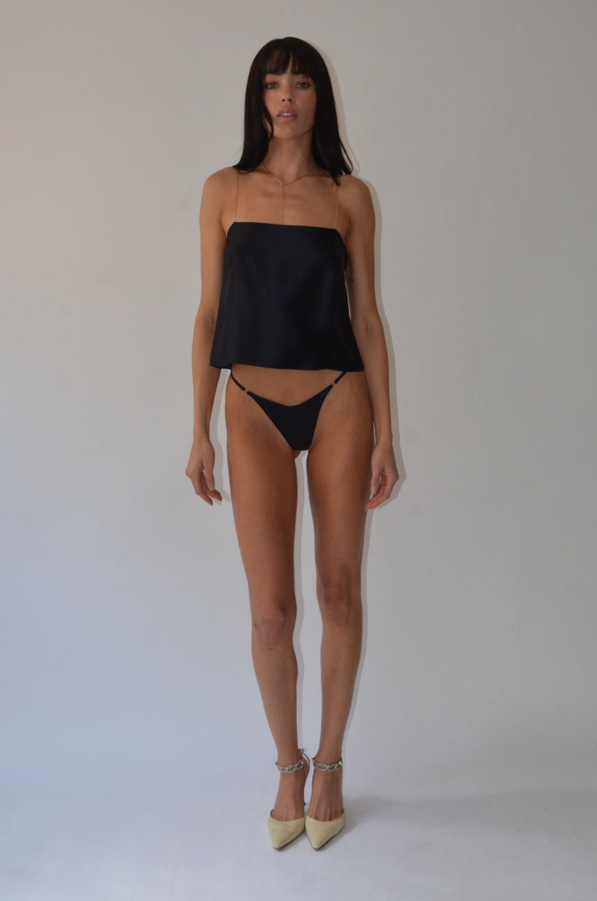 A woman modeling a black strapless tube top and matching black bikini bottoms, paired with beige pointed-toe heels, standing against a plain light gray studio background.
