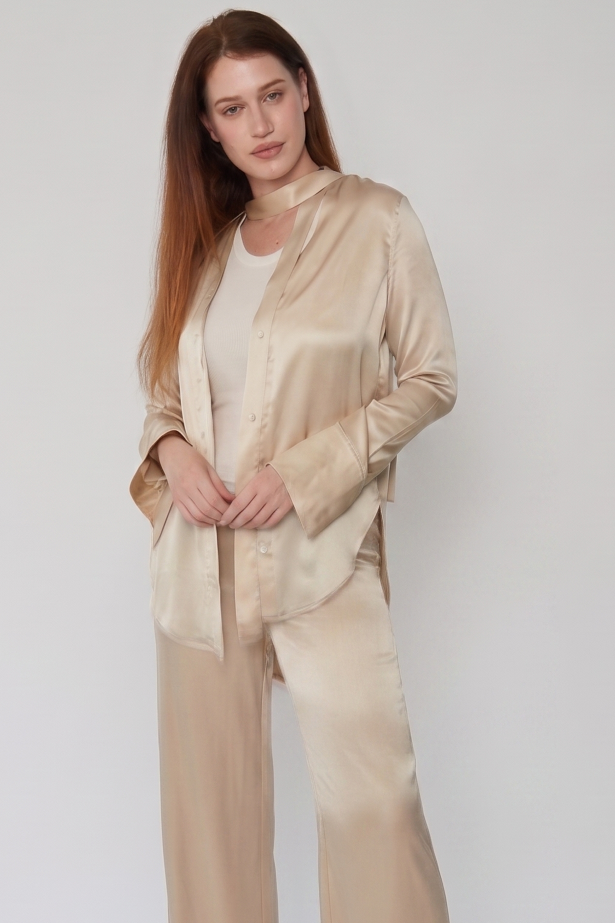 A woman modeling a matching satin pajama set in a light champagne beige color, featuring a long-sleeve button-up shirt and wide-leg pants, worn over a white t-shirt against a plain light gray background.