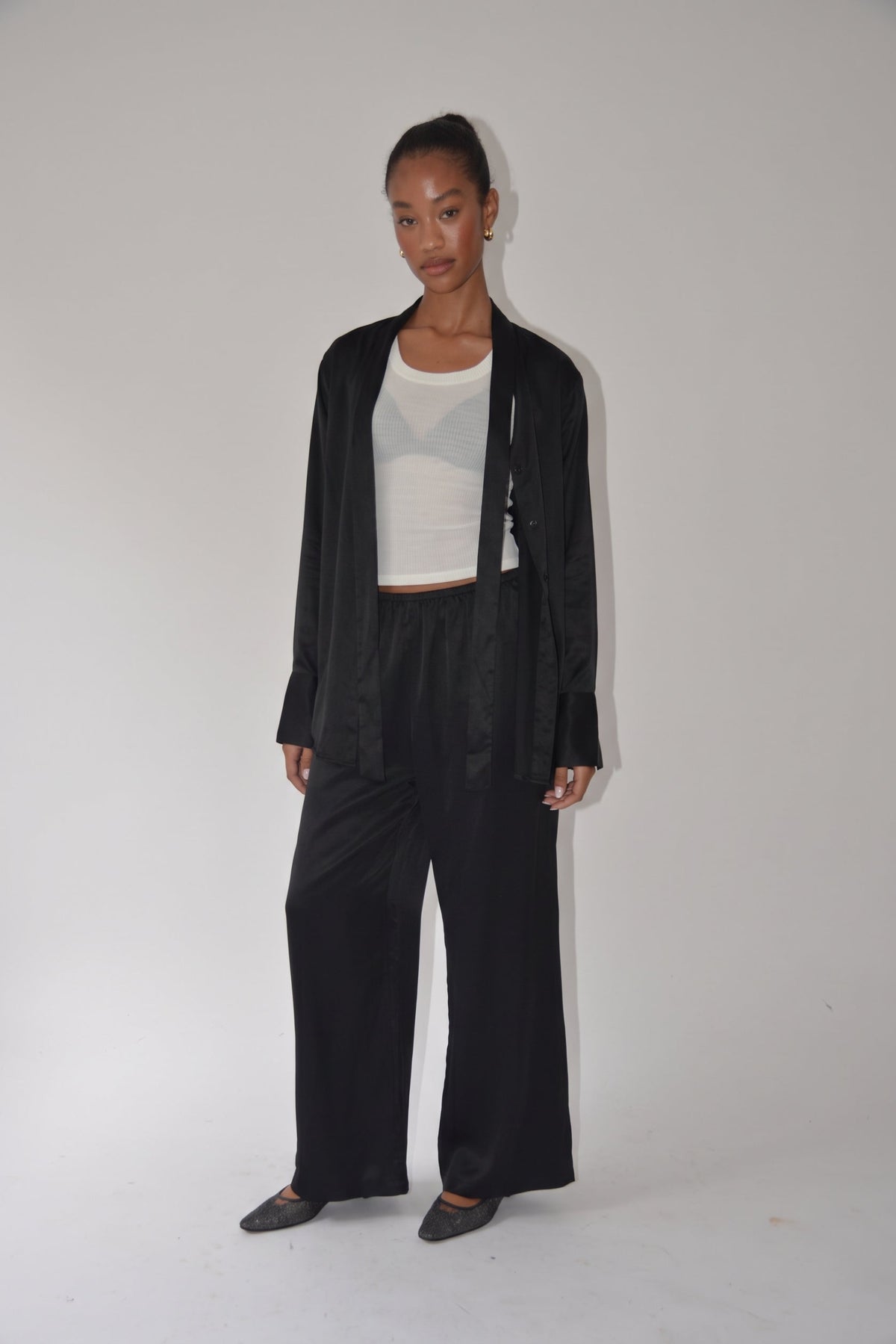 A model stands against a plain light gray studio background wearing a black satin-like open-front jacket, wide-leg black pants, a white cropped tank top, and black pointed flats.