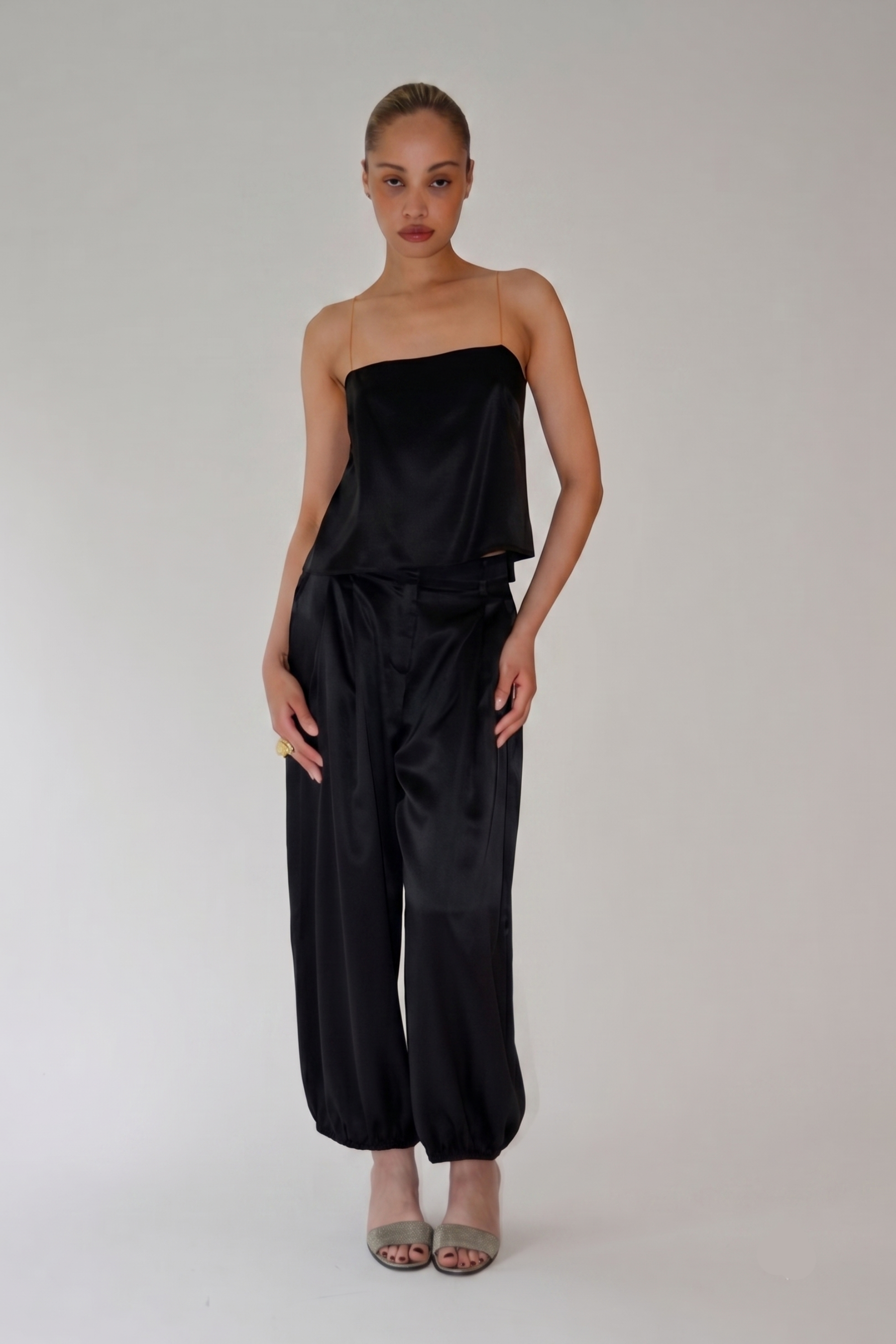 A model stands against a light gray studio background wearing a black satin strapless top and matching black satin wide-leg pants, paired with taupe heeled sandals.
