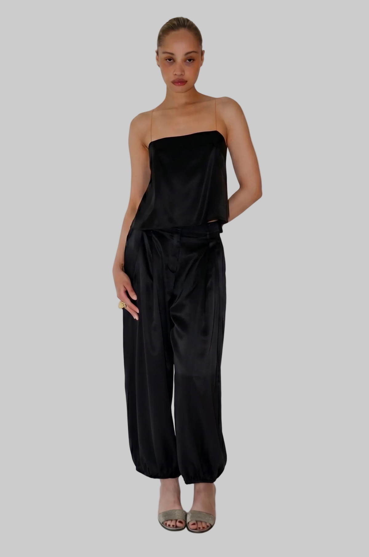 A woman modeling a black satin strapless top and matching wide-leg pants against a plain light gray studio background.
