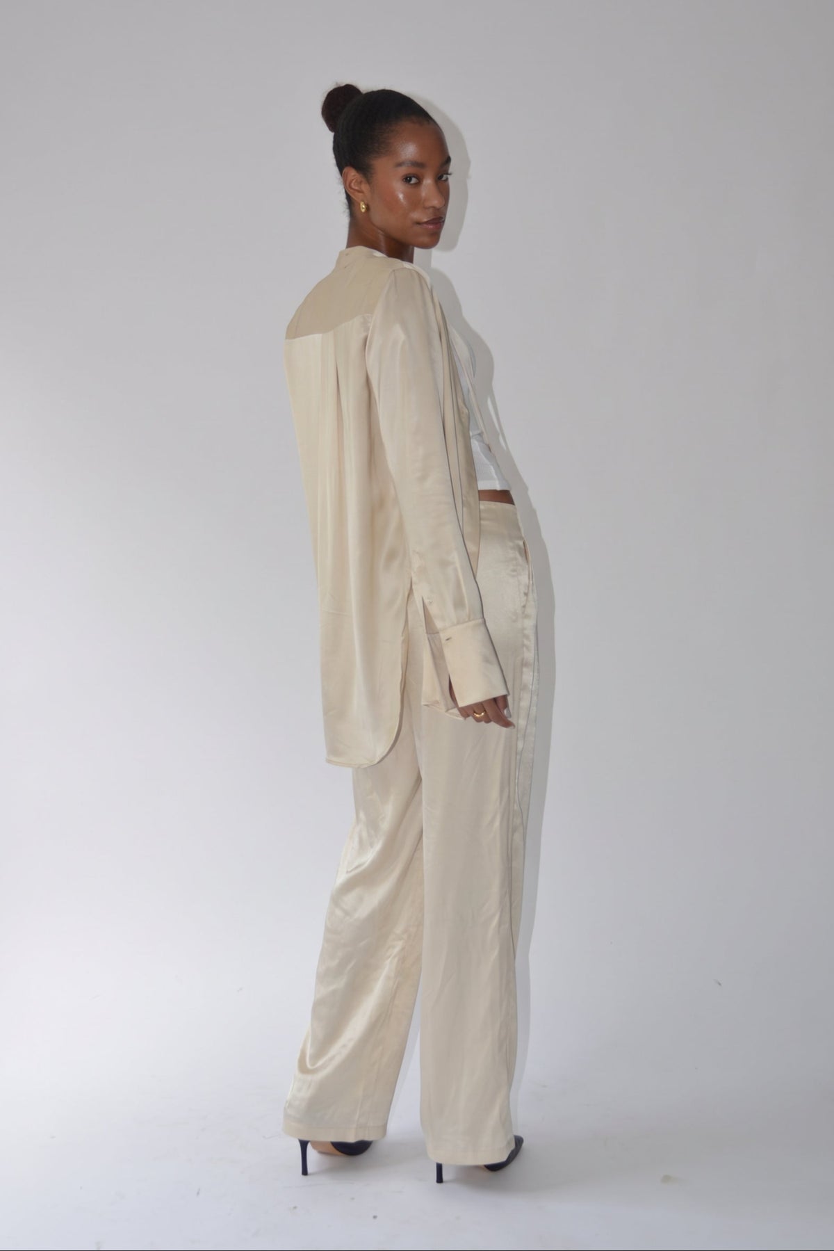 A model is shown from the back wearing a matching cream-colored two-piece suit consisting of a long-sleeve cropped blazer and wide-leg trousers, paired with black high heels against a plain white studio background.