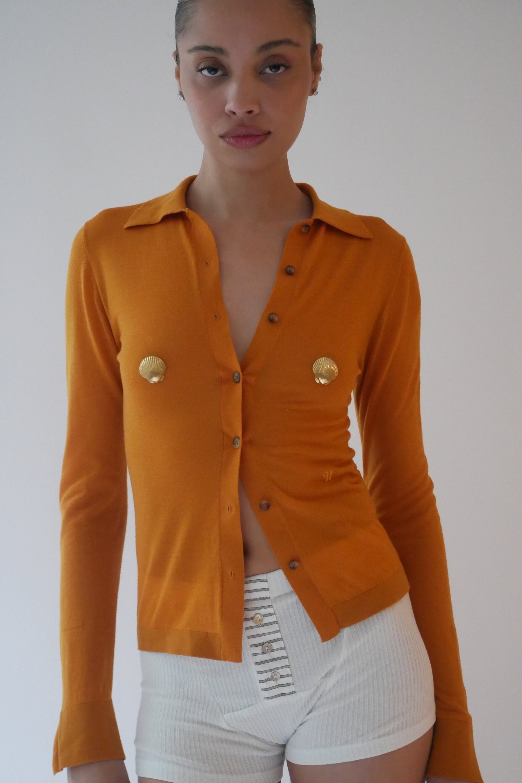 Woman wearing a mustard yellow button-up shirt with gold shell pasties against a plain background