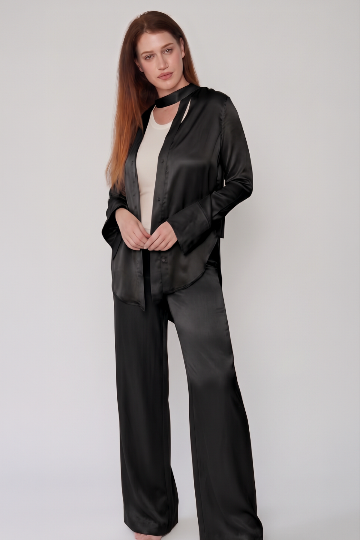 A woman modeling a black satin pajama set consisting of a long-sleeve button-up top with a choker collar and matching wide-leg pants, worn over a cream-colored top, against a light gray studio background.